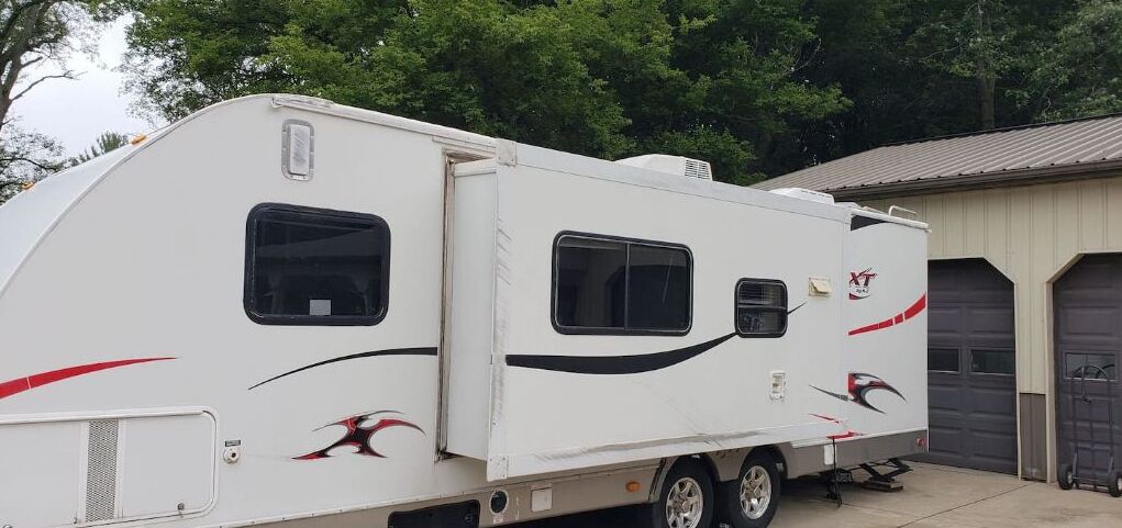 A white RV with two windows and a small door, parked next to a building with two garage doors.