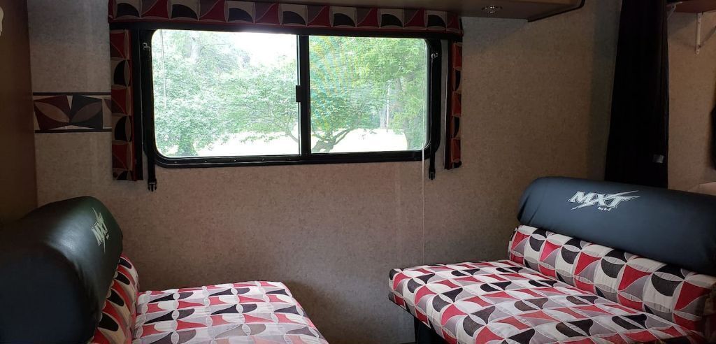 Un intérieur de camping-car compact comprenant deux coins salon, une fenêtre avec des rideaux et un mur décoré d’un motif.