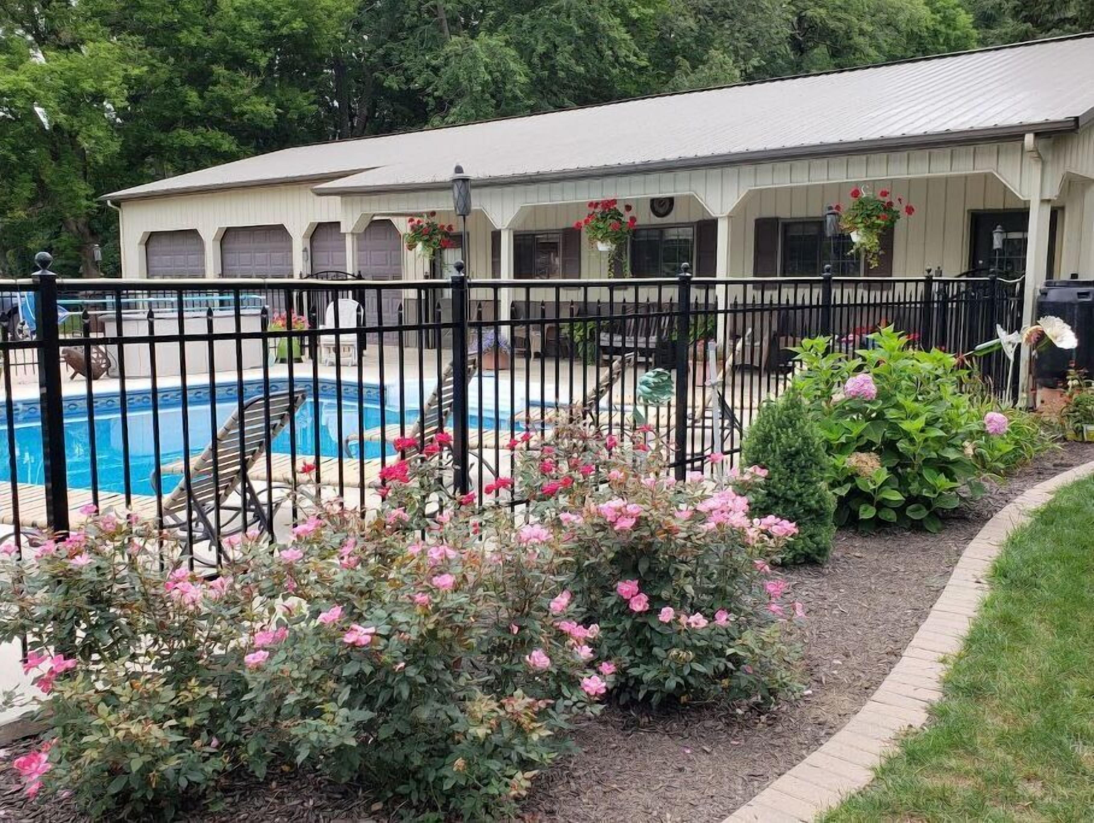 Zona de piscina con terraza cercada, rodeada de flores rosadas y un edificio de fachada blanca.