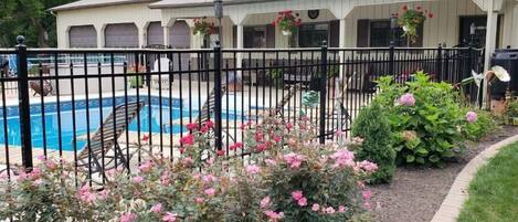 Zona de piscina con terraza cercada, rodeada de flores rosadas y un edificio de fachada blanca.