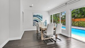A modern dining area with a pool view, featuring a wooden table and white chairs, and a large abstract painting on the wall. - Villa Coastal Cove Sparkling Pool Close to Beach BBQ En-Suite (Boca Raton)
