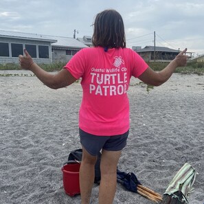 Eine Person mit einem rosa T-Shirt, auf dessen Rückseite 'Coastal Wildlife Club Turtle Patrol' steht, steht auf einem sandigen Strand, in der Nähe befinden sich ein roter Eimer und eine blaue Tasche.