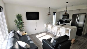 A modern living room with a black leather sofa, a dining area with bar stools, a kitchen with stainless steel appliances, and a large window providing natural light. - Serene 2 bedroom condo with balcony and lakeview plus Free Parking (Winnipeg)