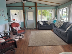 A living room with a grey sofa, wicker chairs, a wooden floor, and a view through a window.