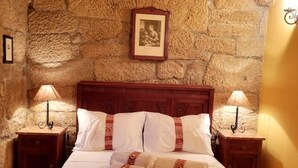 A bed with white linens, two nightstands with lamps, a stone wall, and a framed picture above the bed. - Casa de Campo, Casa do Batocal (Fataunços)