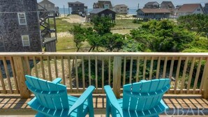Um deck de madeira com duas cadeiras Adirondack azul-turquesa voltadas para uma praia, com casas ao fundo.