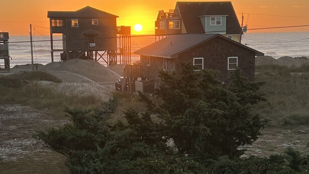 Uma casa à beira-mar com varanda de madeira e um pequeno galpão, com vista para o oceano ao pôr do sol.