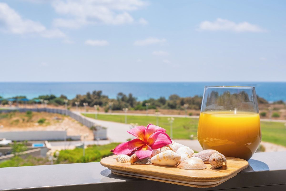 Un verre de jus d’orange sur un plateau en bois, accompagné de quelques coquillages et d’une fleur rose, avec une vue sur un paysage côtier.