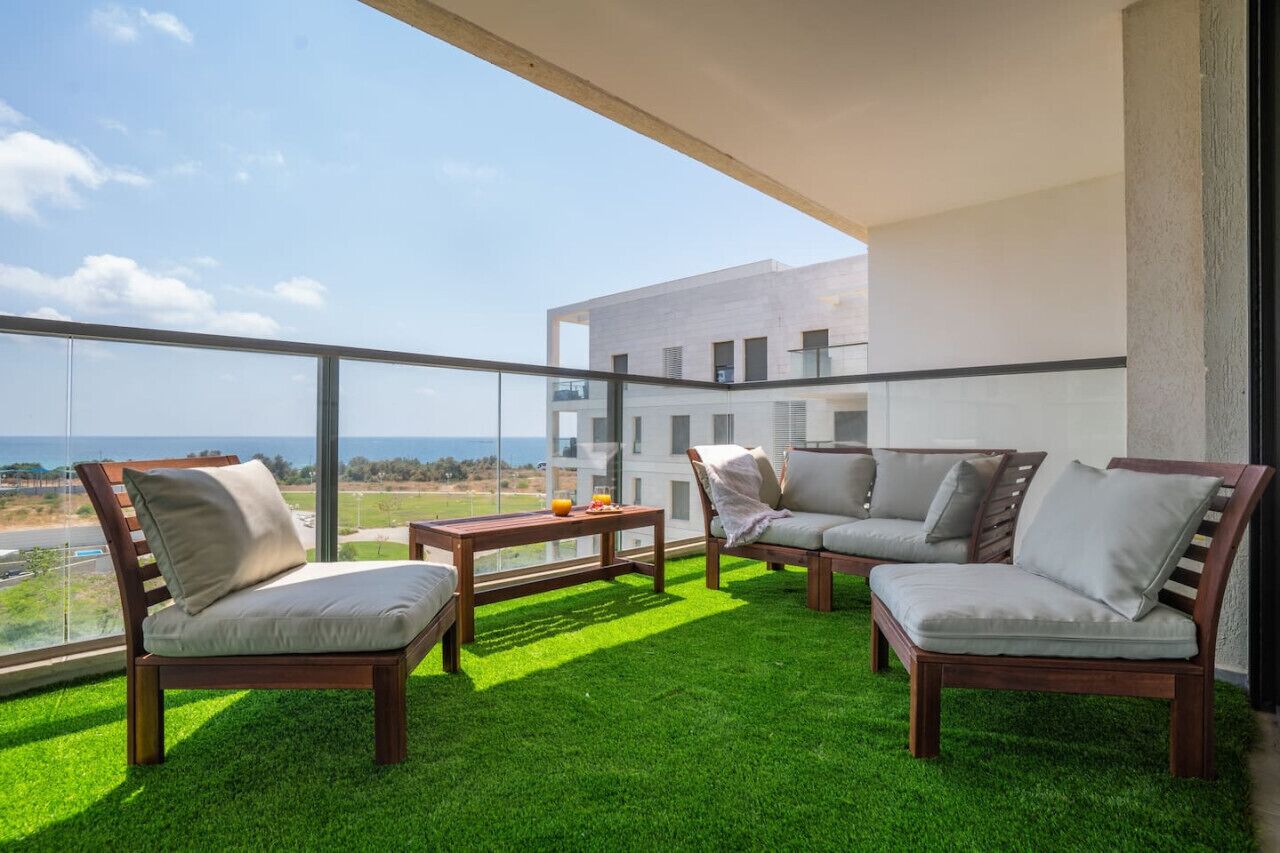 Un balcon doté d’un sol recouvert de gazon artificiel vert, de deux chaises en bois, d’une table en bois et offrant une vue sur un immeuble et l’océan.