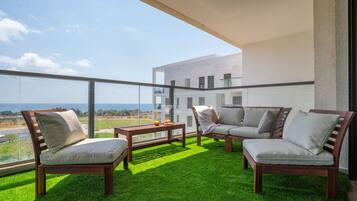 Un balcon doté d’un sol recouvert de gazon artificiel vert, de deux chaises en bois, d’une table en bois et offrant une vue sur un immeuble et l’océan.