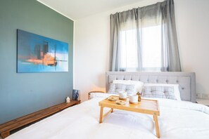 Een slaapkamer met een bed, een nachtkastje, een dienblad met spullen, een schilderij aan de muur en een raam met gordijnen.