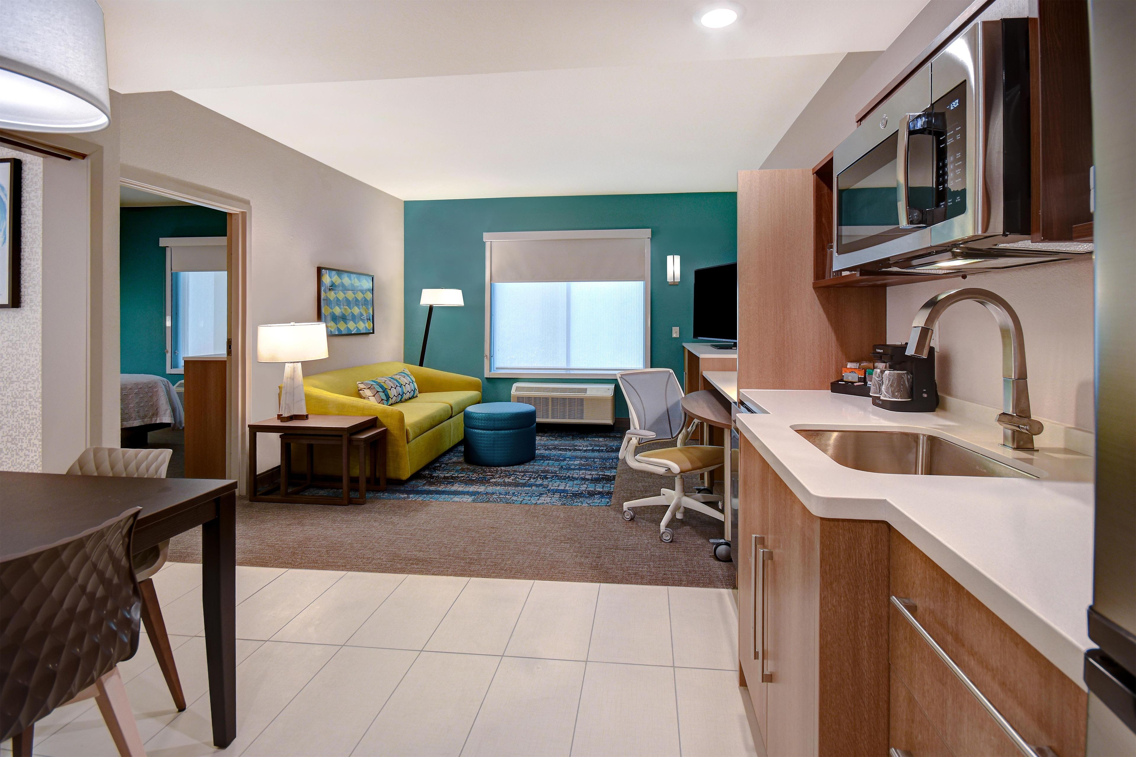 Un espacio habitable compacto con kitchenette, comedor y zona de estar con sofá y reposapiés.