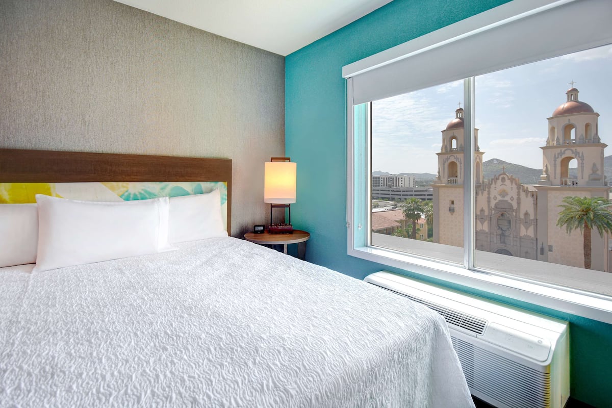 A hotel room with a bed, a nightstand, a lamp, and a window with a view of a church and buildings.