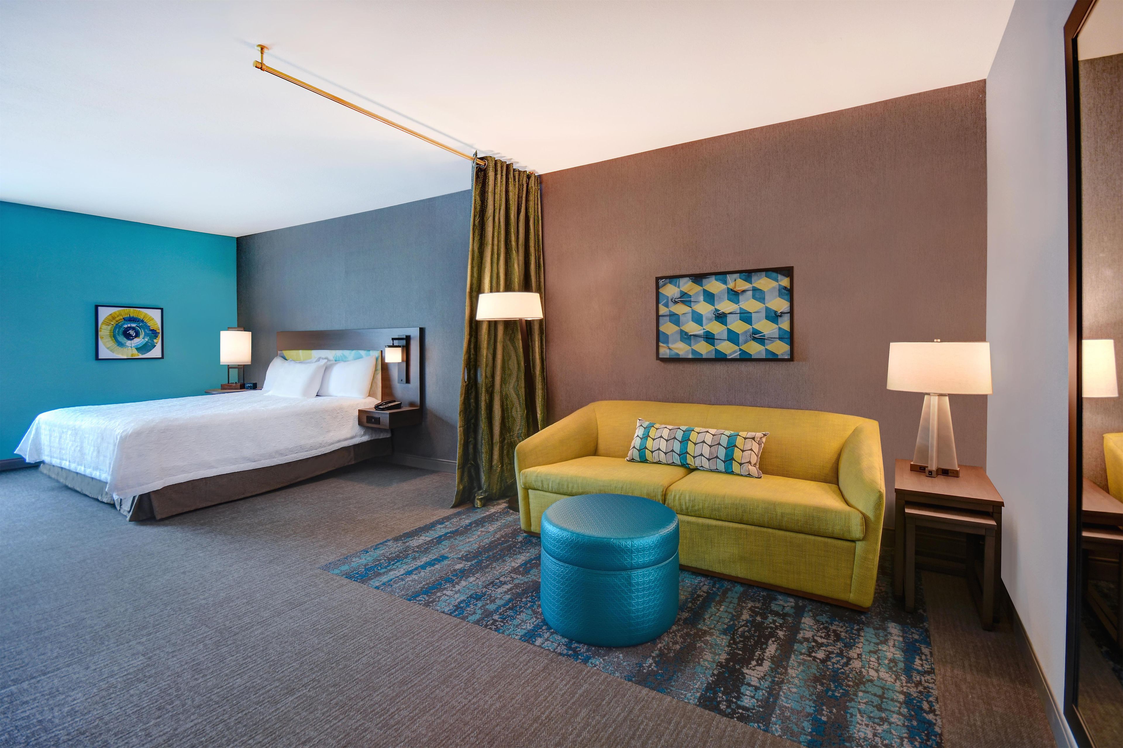 A hotel room with a bed, a sofa, a blue ottoman, a patterned rug, and a wall clock.