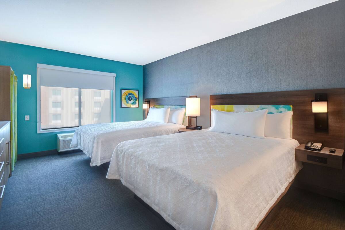 A hotel room with two beds, a window with blinds, a wall-mounted lamp, and a small table with a phone.