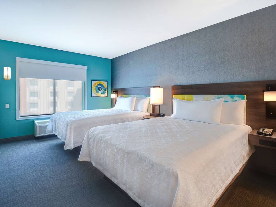 A hotel room with two beds, a window with blinds, a wall-mounted lamp, and a small table with a phone.