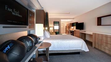 Una habitación de hotel con cama, escritorio, equipo de ejercicio, televisor y espejo.