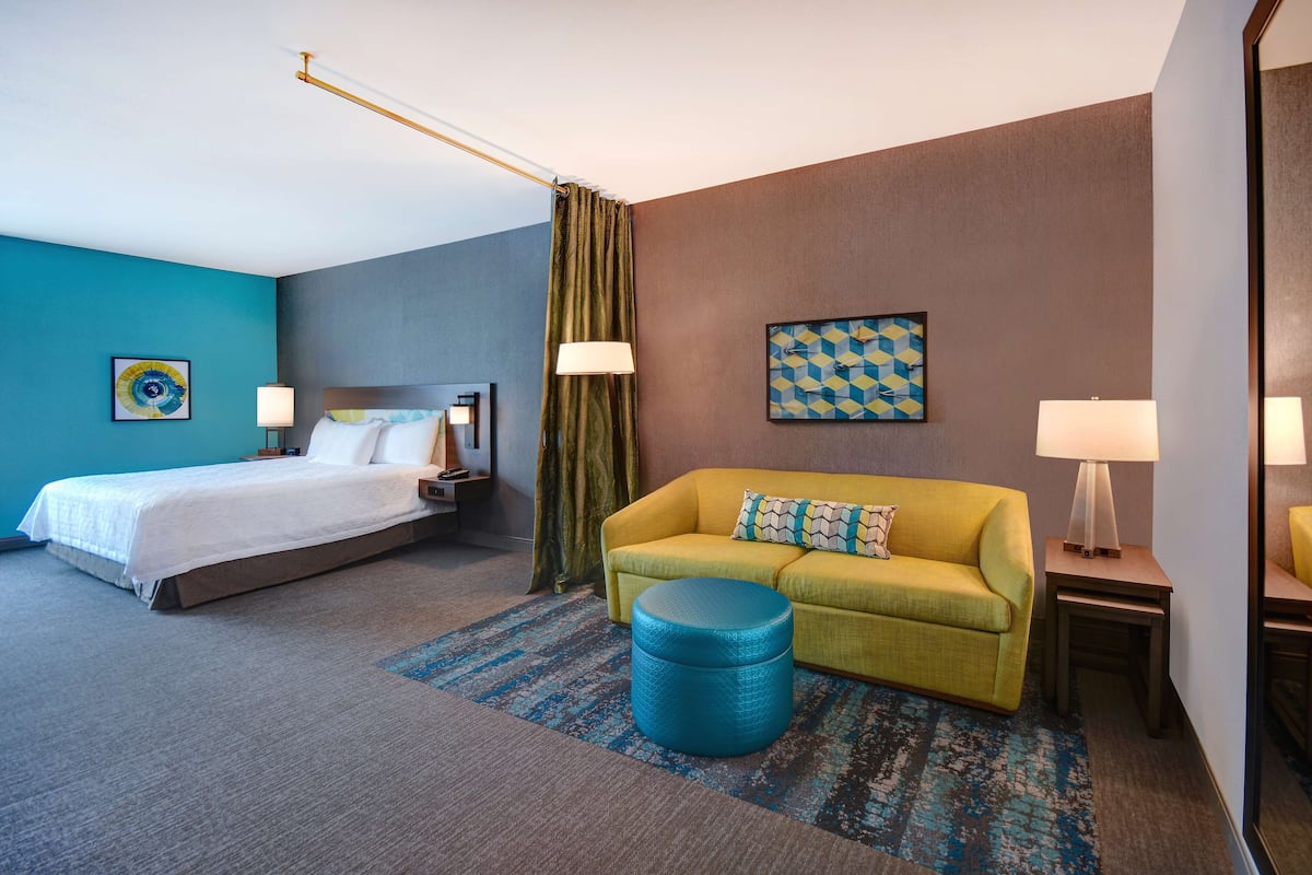 A hotel room with a bed, a yellow sofa, a blue ottoman, and a blue and brown patterned rug.