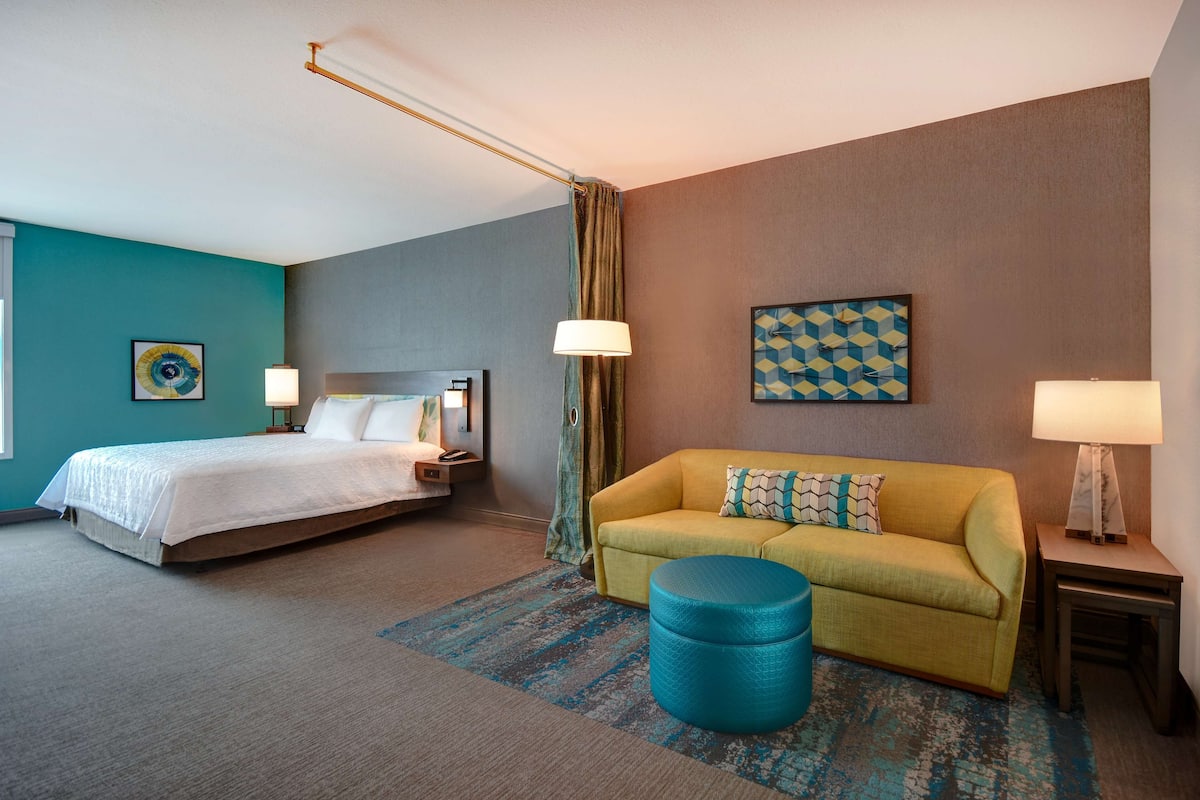 A hotel room with a bed, a sofa, a blue ottoman, and two lamps.