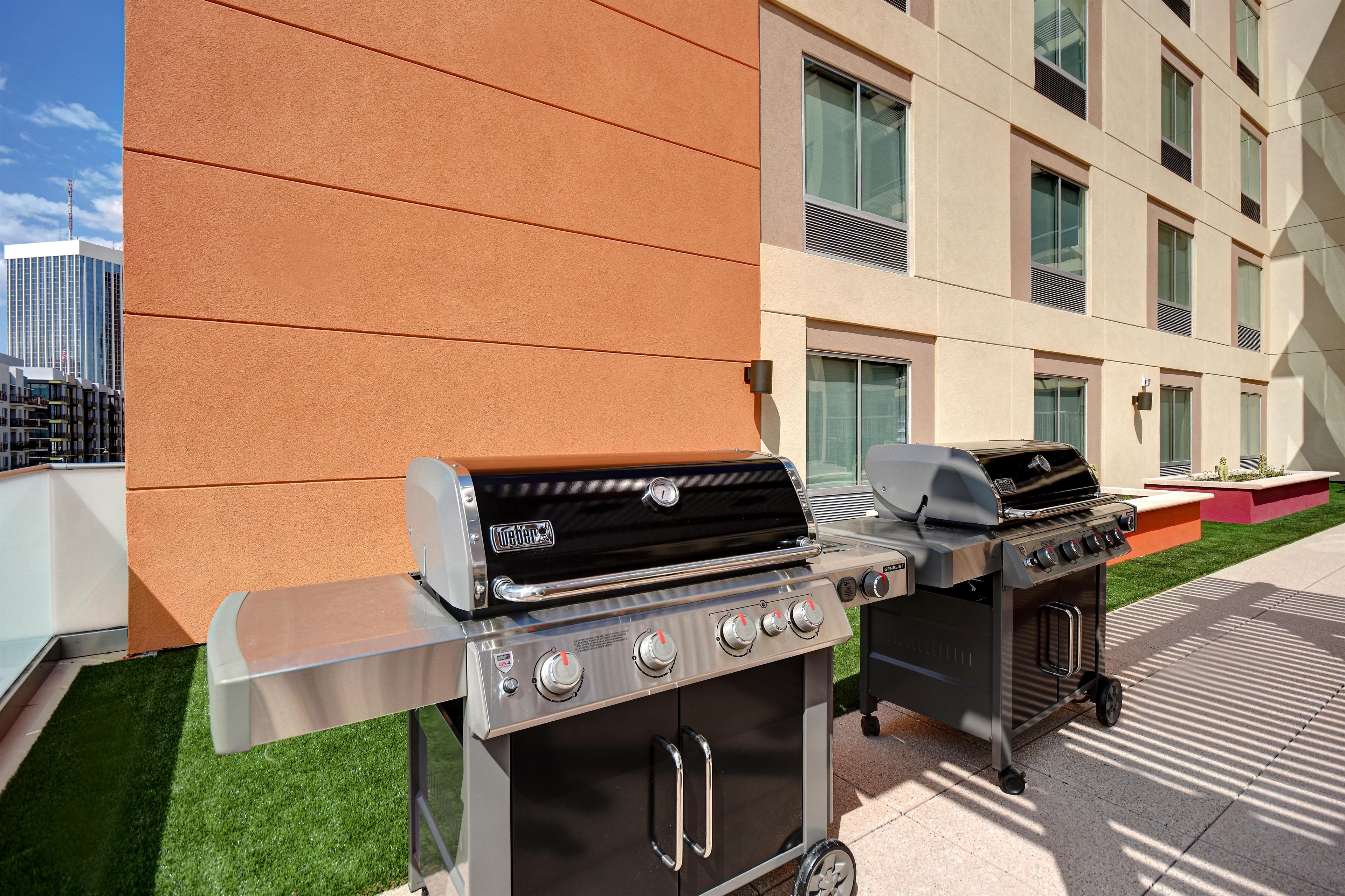 An outdoor grill area with a Weber brand grill and a portable gas grill, set against a modern building facade.