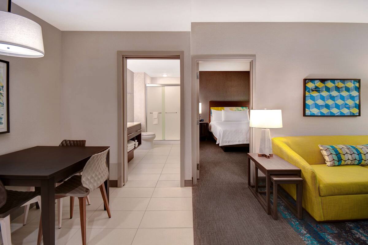 A hotel room with a dining table, chairs, a yellow sofa, a bed, and a bathroom visible through an open door.