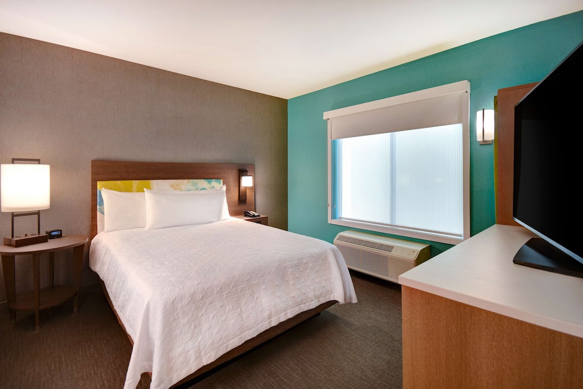 A hotel room with a bed, bedside table with lamp, TV, and window with blinds.