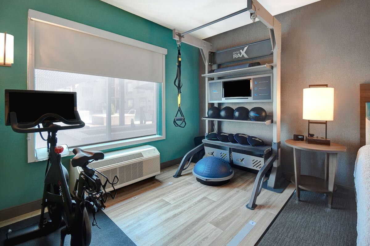 A compact gym with a stationary bike, a pull-up bar, a medicine ball, and a rack with exercise balls and dumbbells.