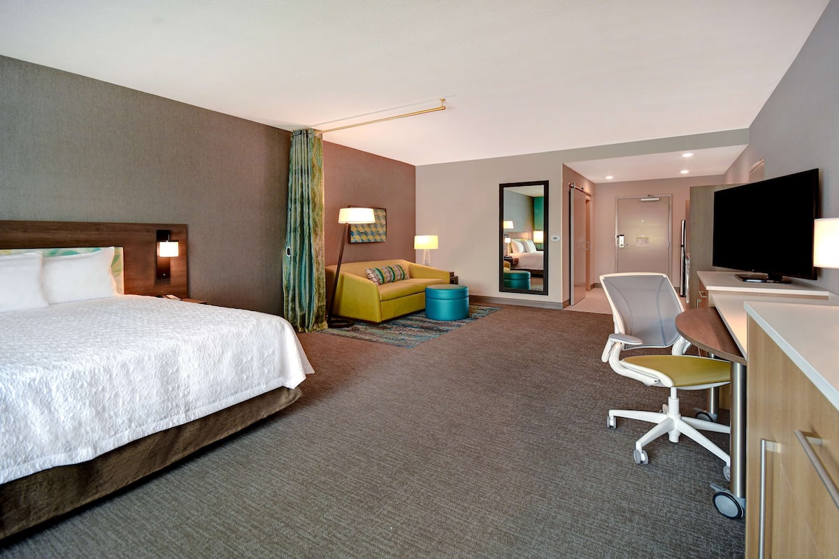 A hotel room with a large bed, a sofa, a desk with a chair, a television, and a bathroom visible through an open door.