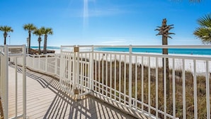 A wooden deck with a white railing overlooking a beach with palm trees and clear blue sky.