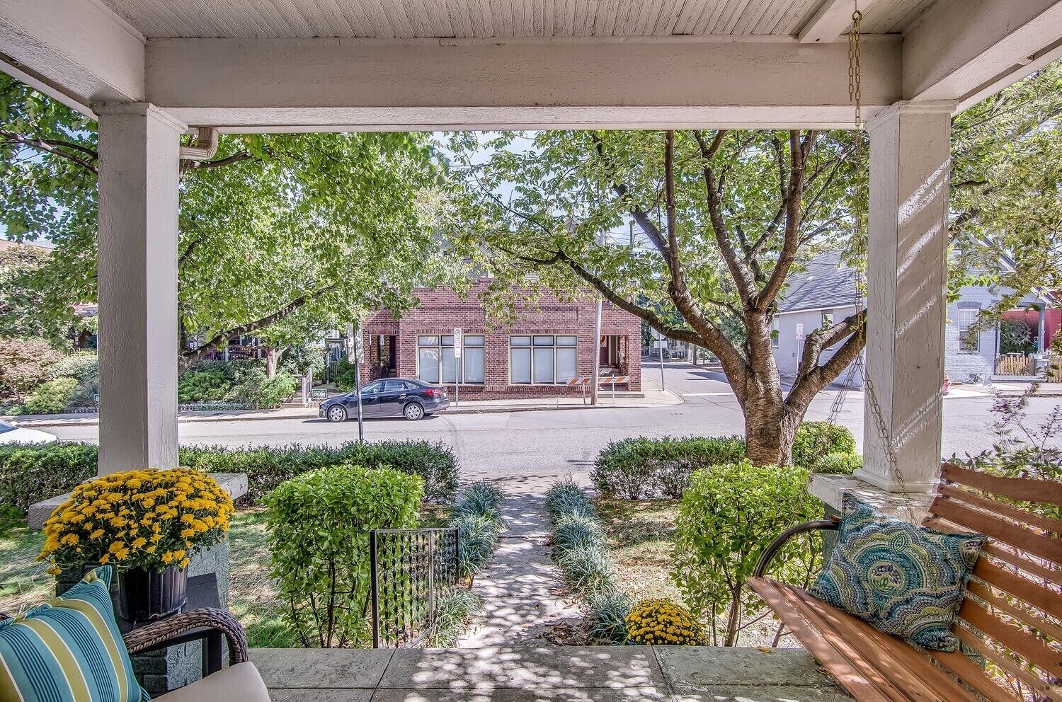 A covered porch with a view of a brick building, a car on the street, and a bench with a patterned cushion.