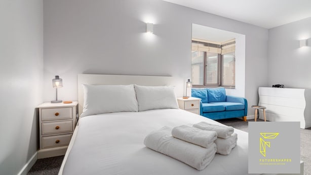 Una habitación de hotel moderna con una cama grande, un sofá azul, un tocador blanco y una mesita de noche con una lámpara.