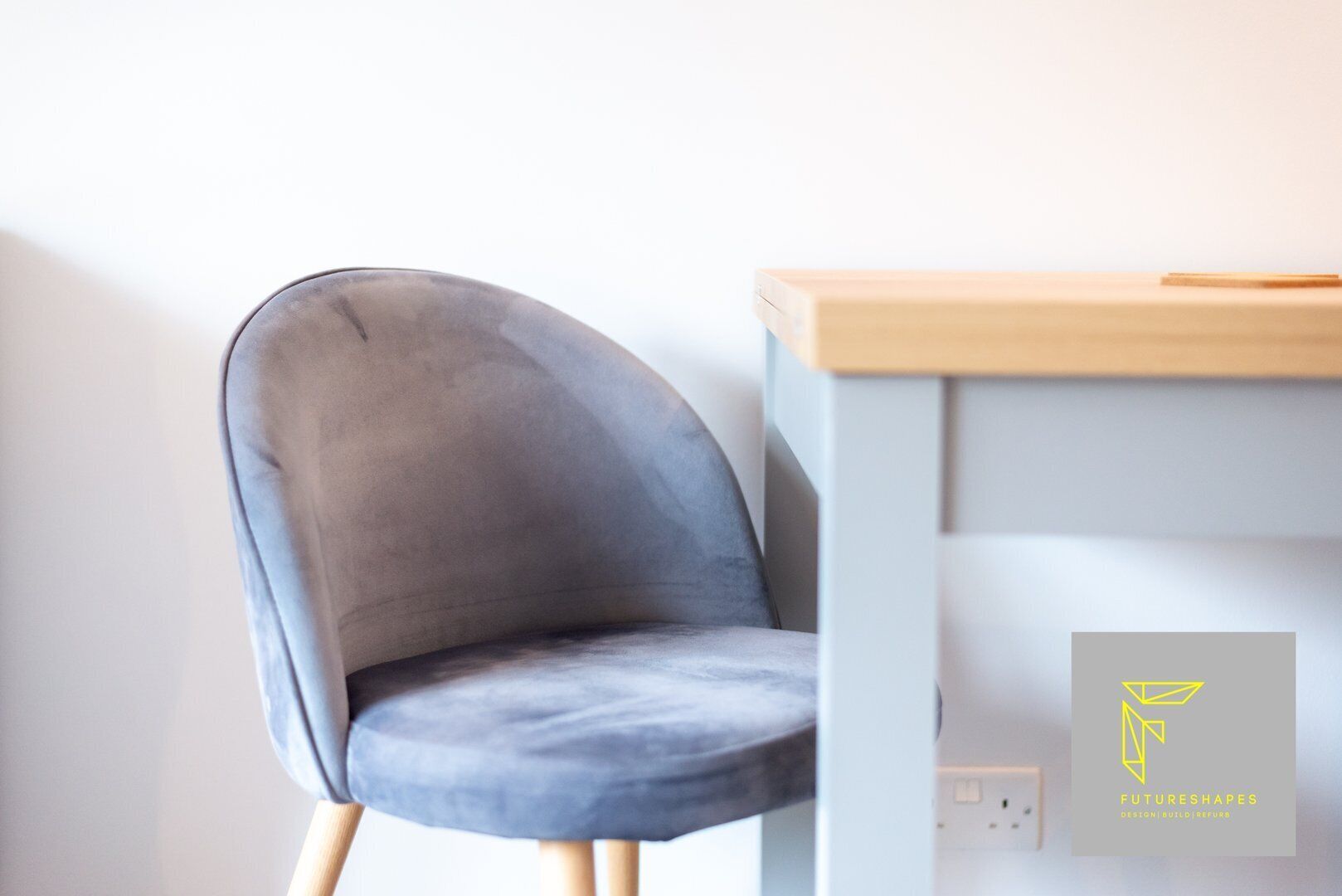 A modern chair with a smooth, curved backrest and wooden legs, positioned next to a light-colored desk with a wooden top and a drawer.