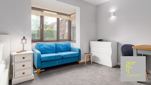 Una habitación moderna y funcional con un sofá azul, un aparador blanco, una mesita redonda pequeña y un escritorio con una silla.