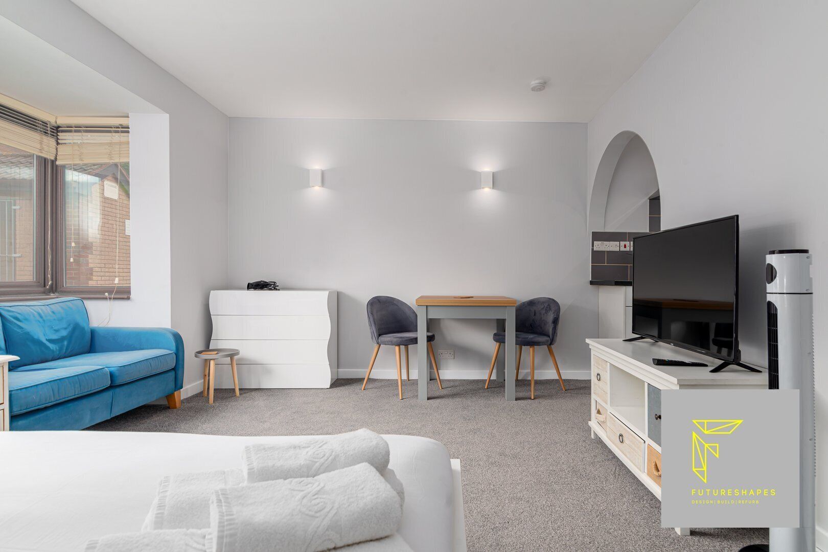 A modern hotel room with a bed, a blue sofa, a desk with two chairs, a TV, and a wardrobe.