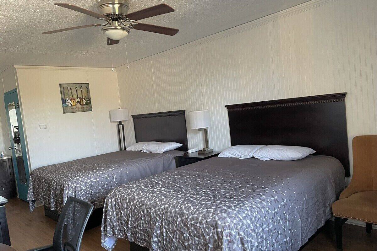 A hotel room with two beds, a ceiling fan, a desk with a chair, and a nightstand with a lamp.