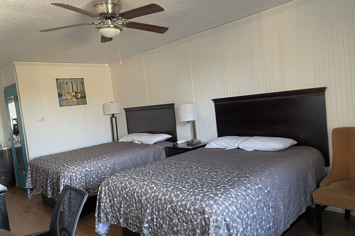 A hotel room with two beds, a ceiling fan, a desk with a chair, and a nightstand with a lamp.