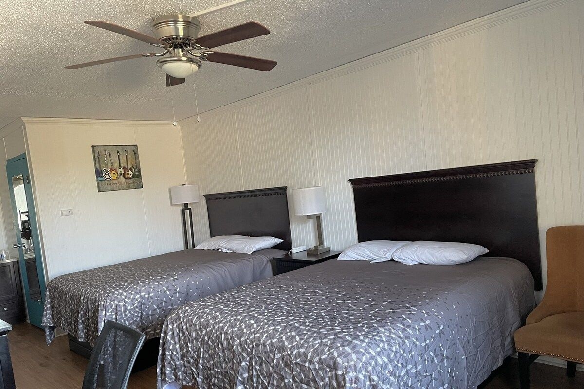 A hotel room with two beds, a ceiling fan, a desk with a chair, and a nightstand with a lamp.