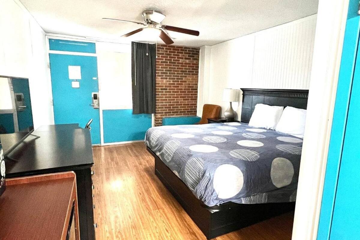 A hotel room with a bed, a desk, a chair, a ceiling fan, and a brick wall.