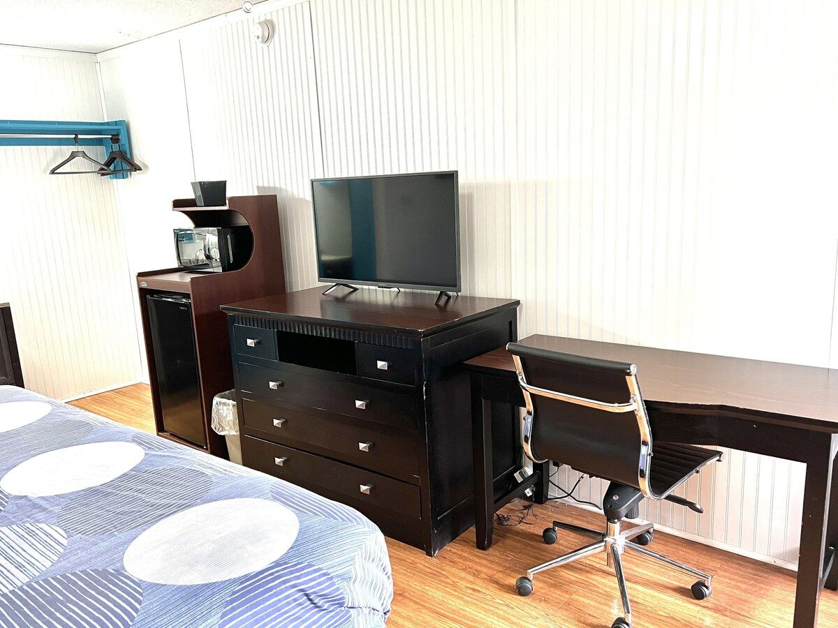 A hotel room with a bed, a desk with a chair, a television, a dresser, and a small refrigerator.