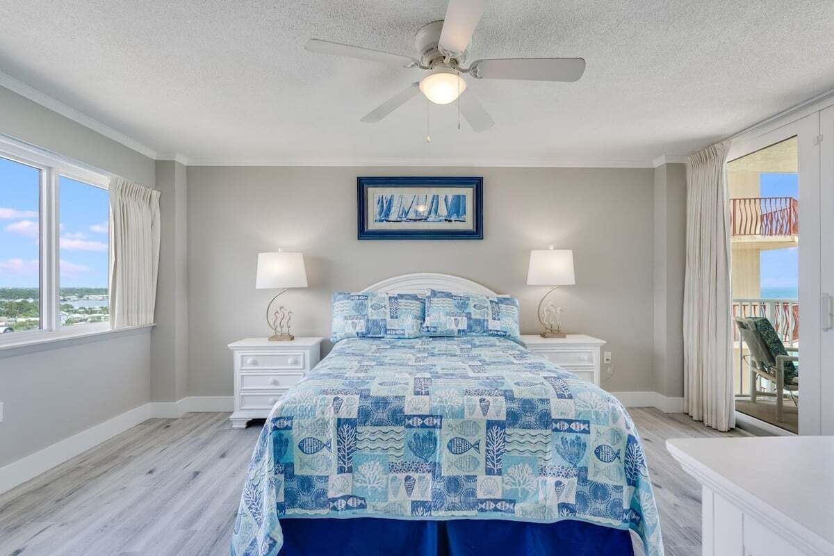 Un dormitorio con cama, mesitas de noche, ventilador de techo, ventana con cortinas y balcón con vistas.