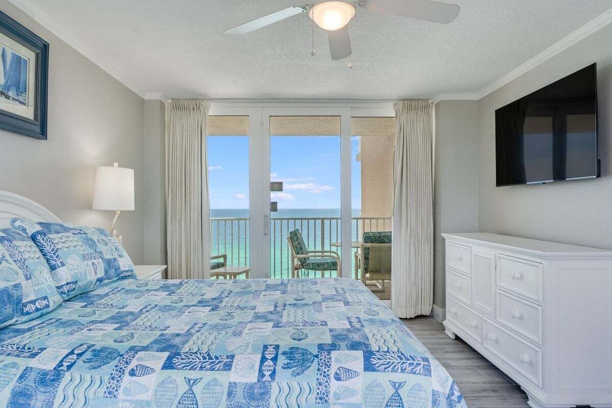 Un dormitorio con cama, ventilador de techo, televisor, balcón con vista al océano y cómoda blanca.