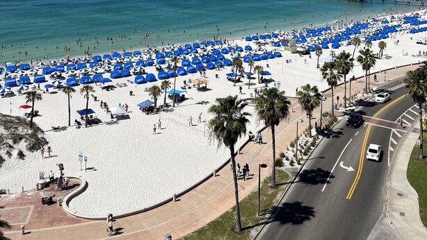 A beach with numerous blue umbrellas and white tents, a road adjacent to the beach, and a pier in the background. - Excellent home for multiple families.  Enjoy the pool or fish off the dock. (Clearwater)