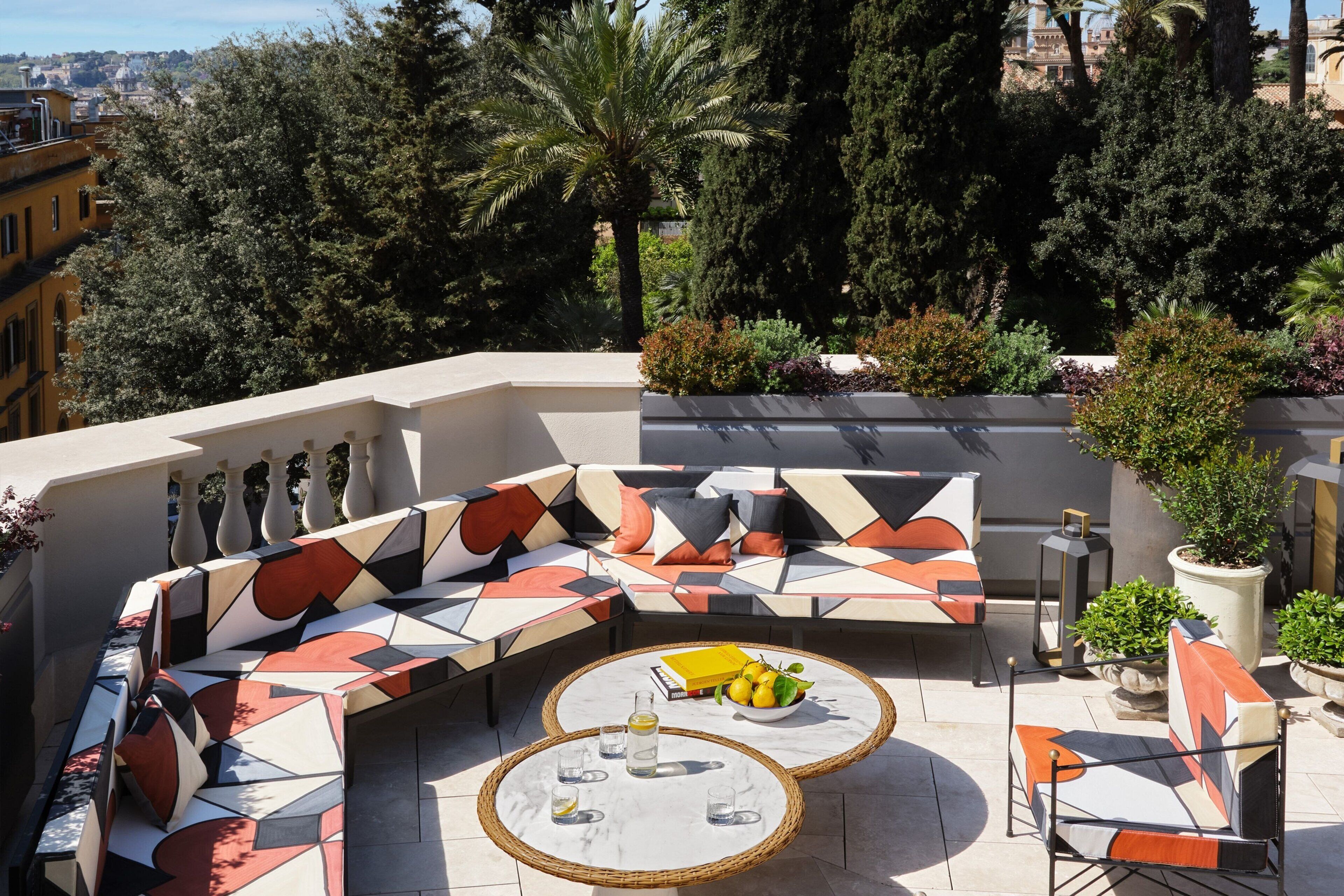 Un espace extérieur moderne, aménagé avec un canapé aux motifs géométriques et deux tables rondes, entouré de plantes en pot et offrant une vue sur le paysage urbain.