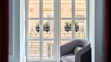 Une pièce dotée d’une grande fenêtre, d’un fauteuil gris et offrant une vue sur un immeuble avec des balcons.