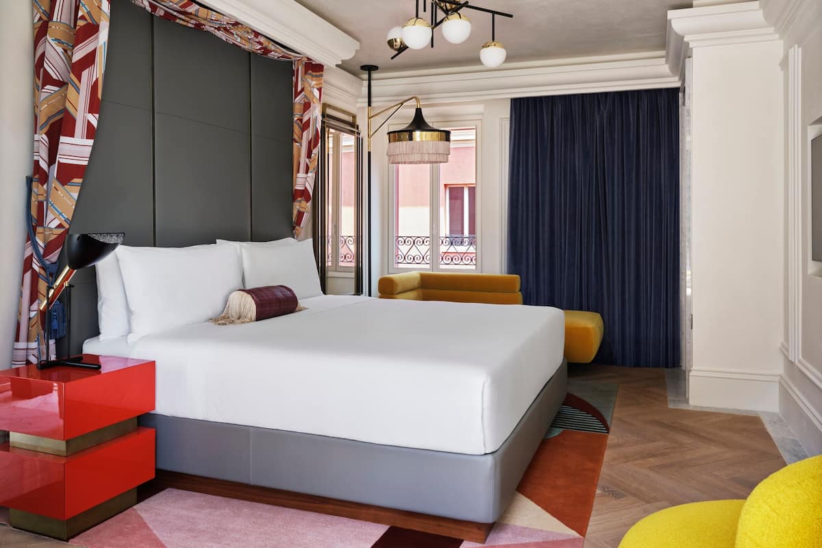 A modern hotel room with a large bed, a red nightstand, a yellow armchair, and a balcony with a view.