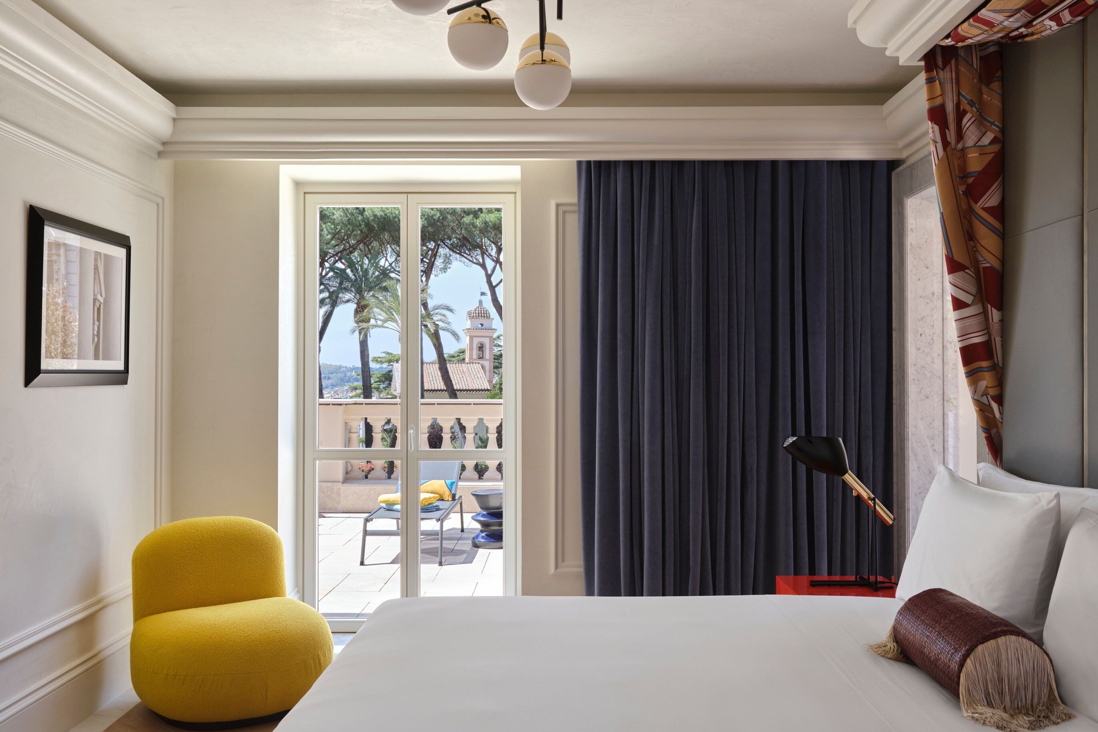 Ein Hotelzimmer mit einem Bett, einem gelben Sessel, einem großen Fenster mit Blick auf Palmen und ein Gebäude, sowie einem dunklen Vorhang.