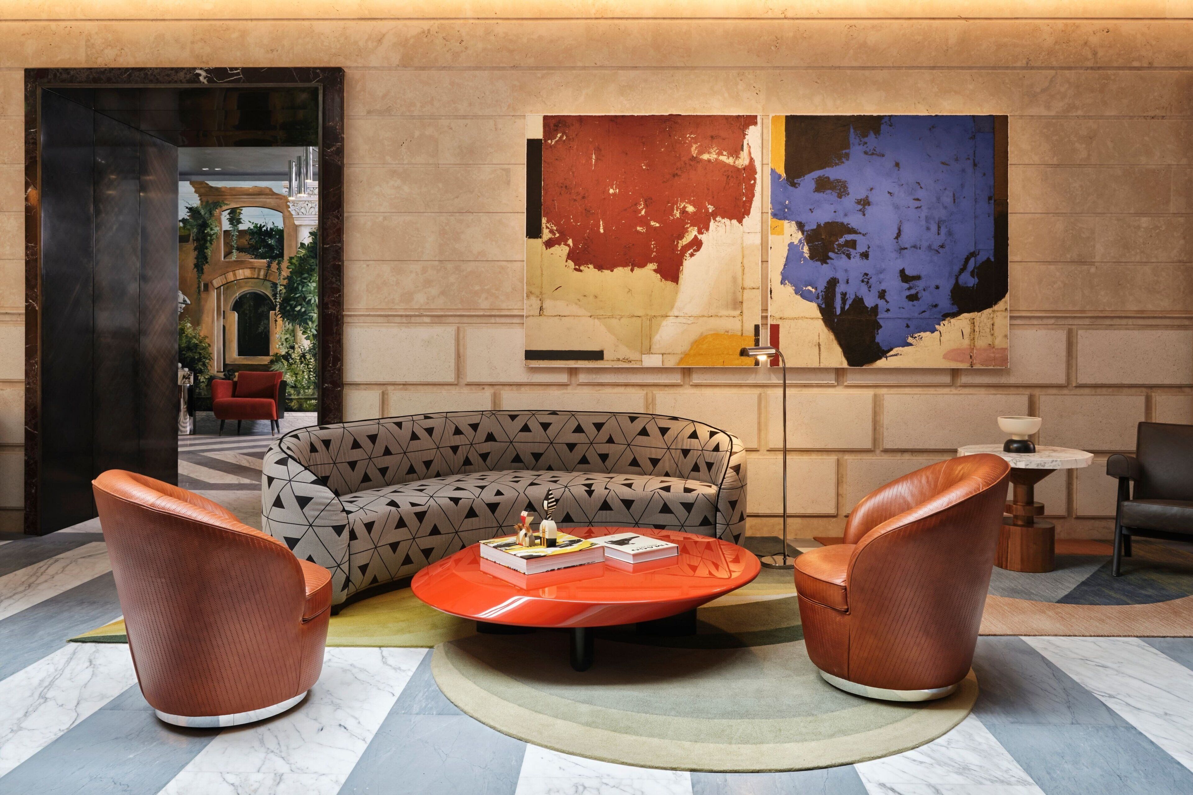 A modern lobby with a patterned sofa, two leather armchairs, a red coffee table, and a large abstract painting on the wall.