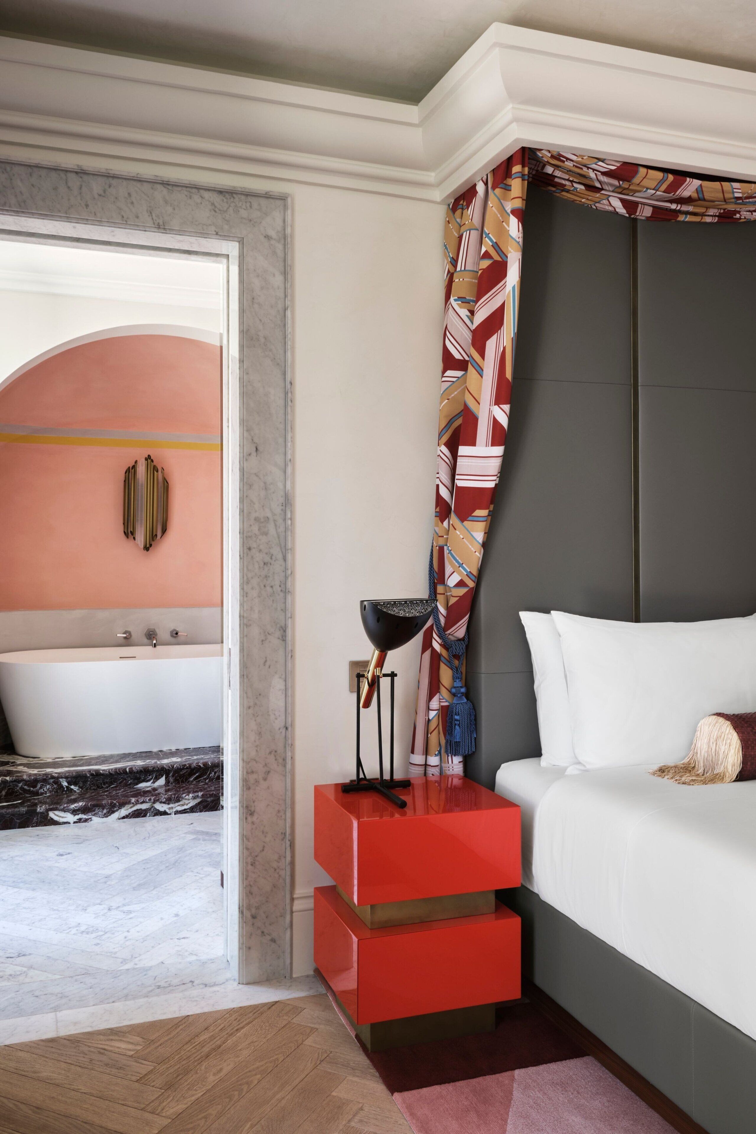 A modern bedroom with a bed, a nightstand, a red drawer unit, a black floor lamp, and a bathroom with a bathtub and a towel rack.