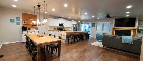 Una cocina moderna con una isla central, comedor con sillas, un sofá y un televisor sobre la chimenea.