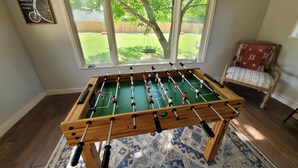 Uma mesa de futebolito numa sala com uma janela grande, uma cadeira e uma frase motivacional na parede.